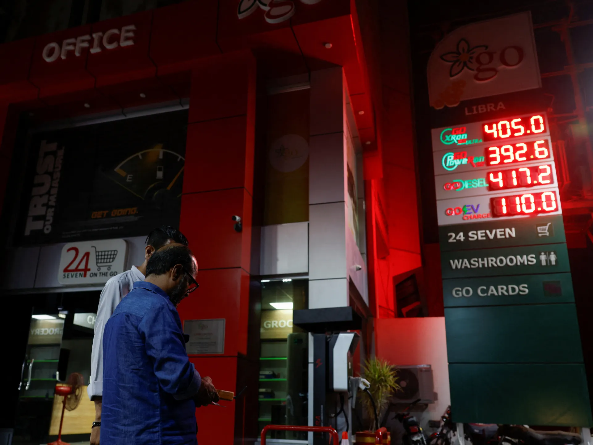 Two men stand beside a fuel station price board glowing red at night.