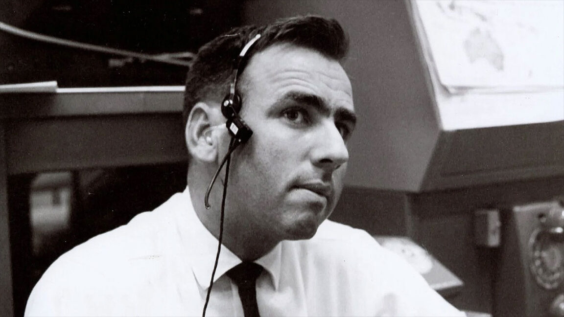 Black-and-white photo of a man wearing a headset, white shirt, and dark tie at a control console.