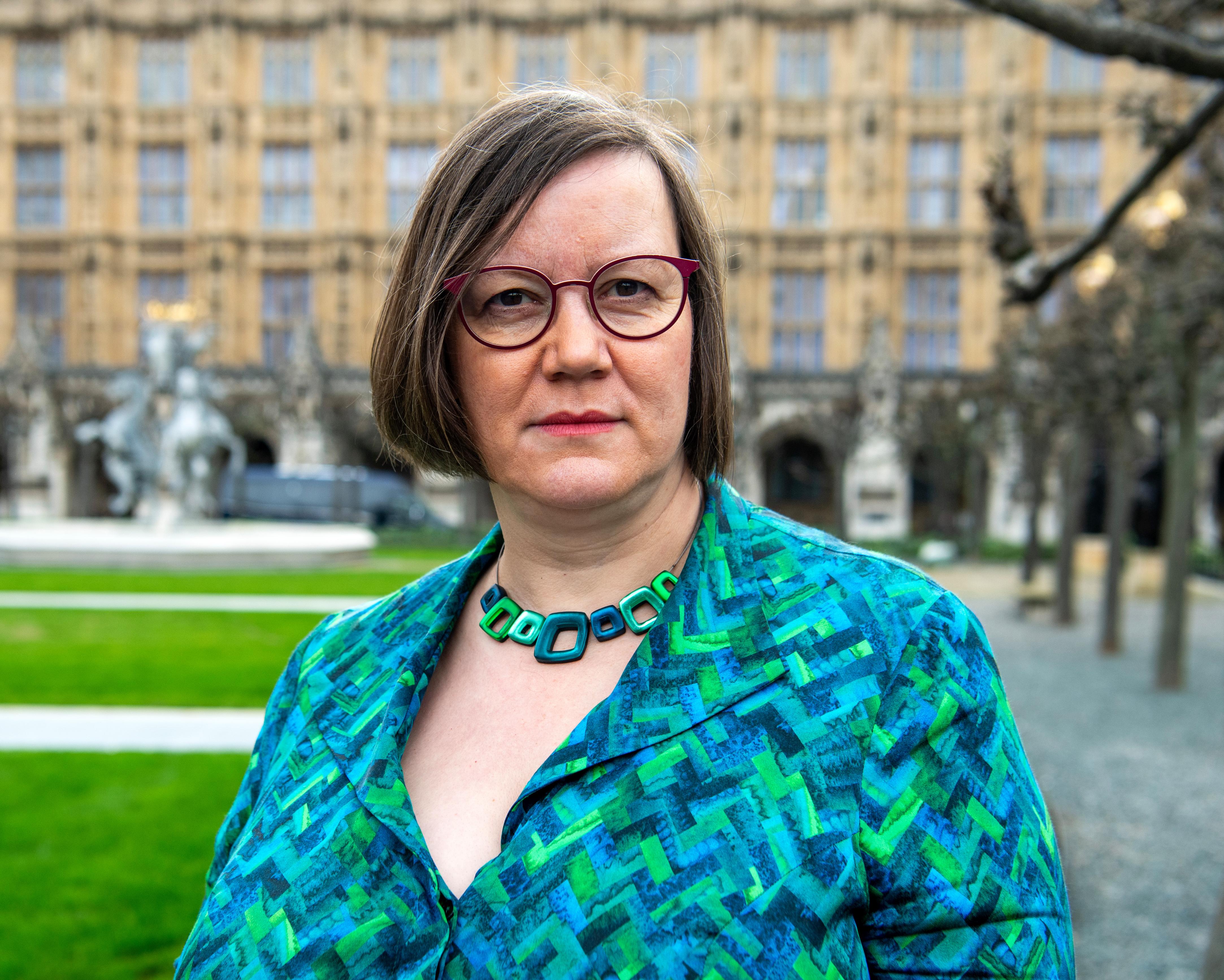 A woman wearing red glasses and a patterned blue-green jacket stands outdoors before a stone building.