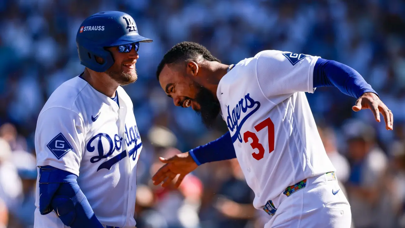 Two Dodgers players in white uniforms smile and gesture toward each other on the field.