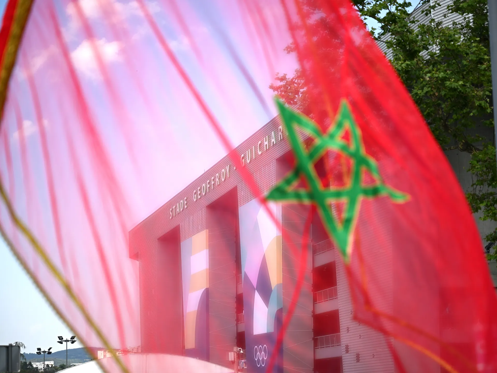Moroccan flag in the foreground partially veils a building facade bearing Olympic rings and colorful banners.