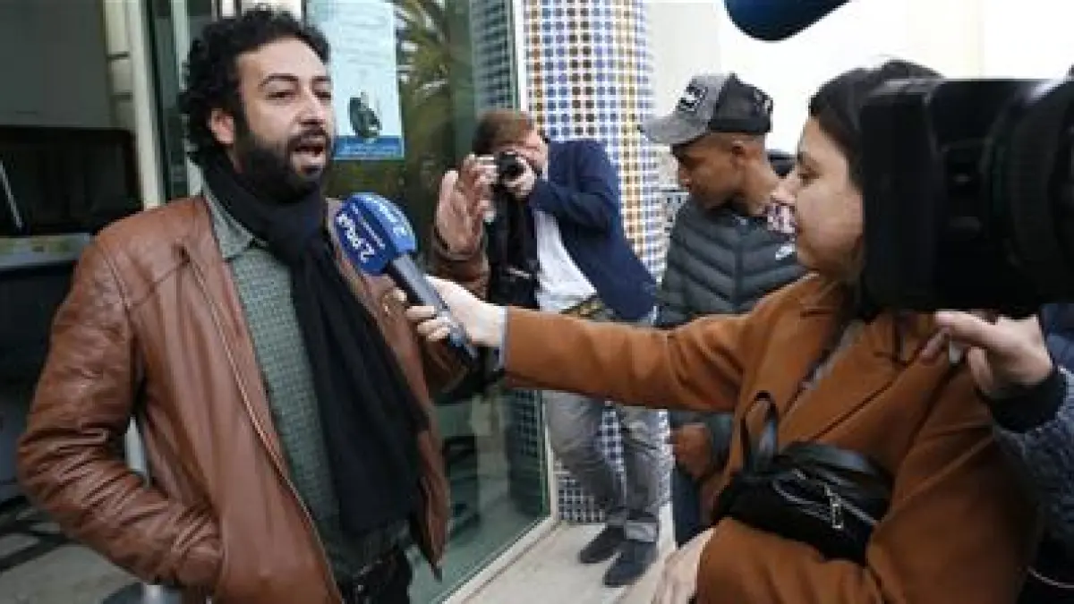 A bearded man in a brown leather jacket speaks into a reporter’s microphone as cameras record nearby.