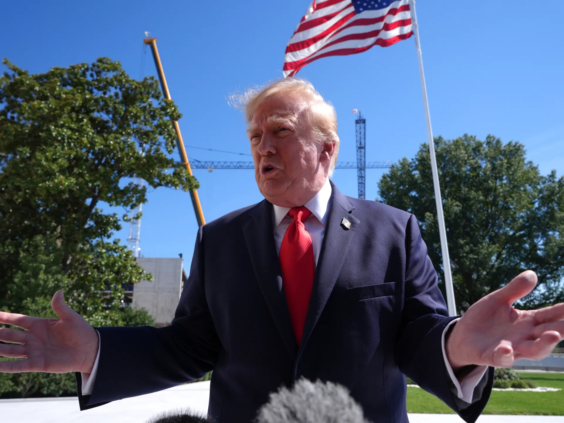 Donald Trump speaks outdoors with arms outstretched beneath a large American flag.