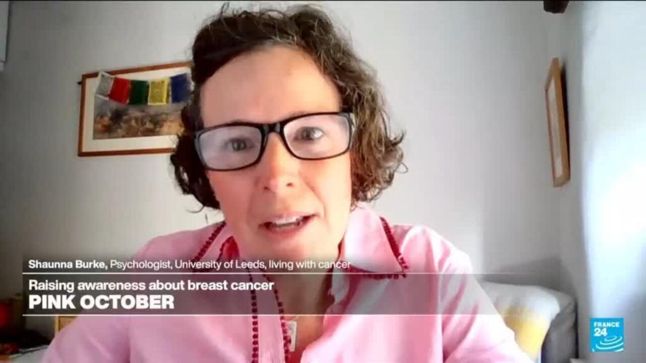 Woman wearing glasses and a pink shirt speaks to camera in a home setting.