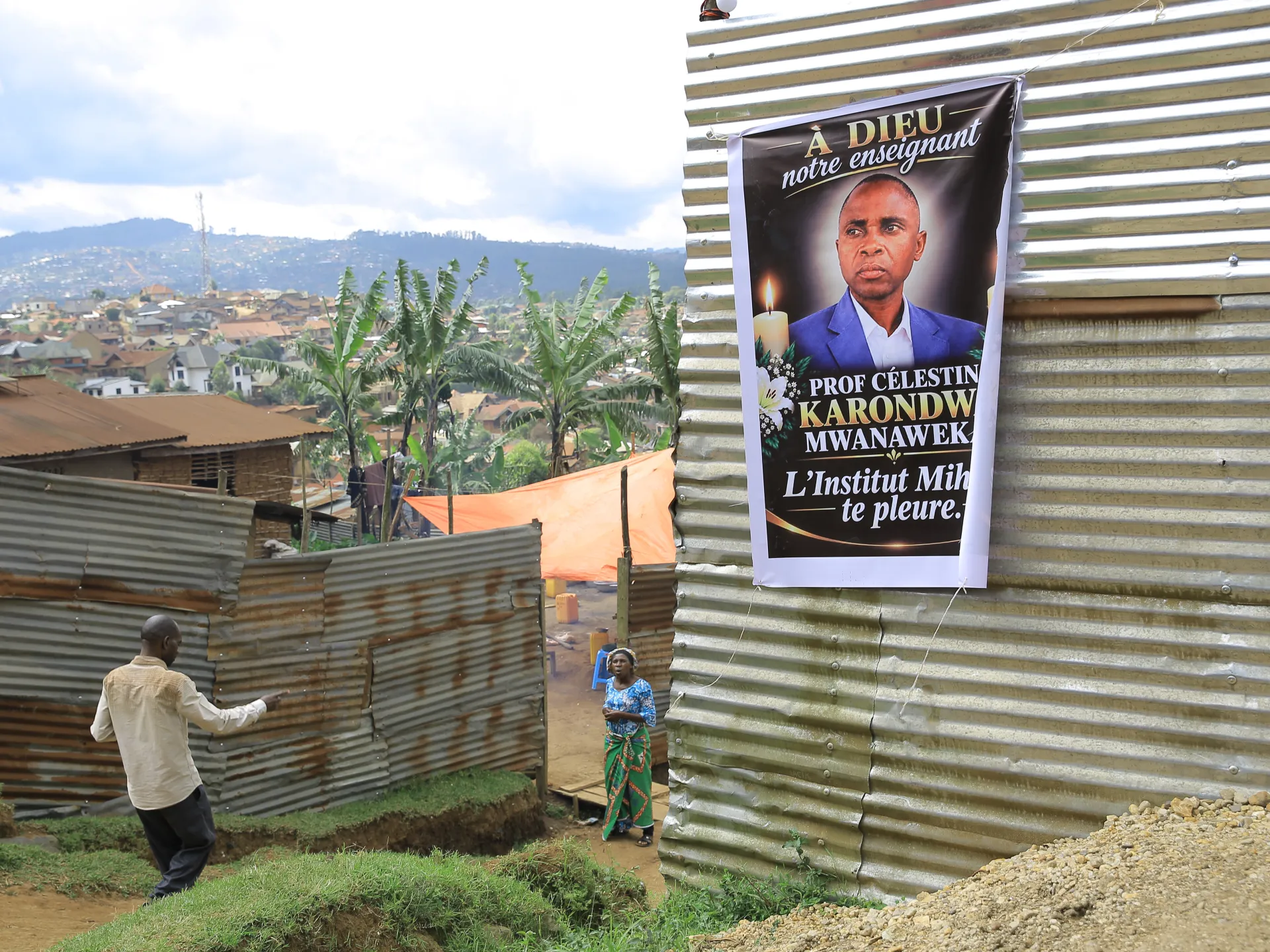 A memorial poster hangs on a corrugated metal wall above a hillside neighborhood, with two people nearby.