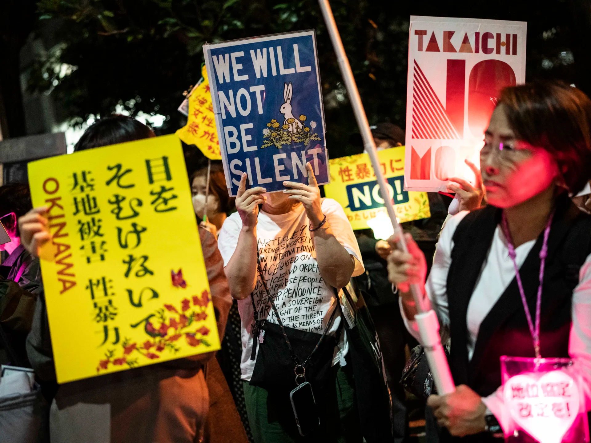 Demonstrators hold protest signs at night, including messages reading “We Will Not Be Silent” and “Takeuchi.”