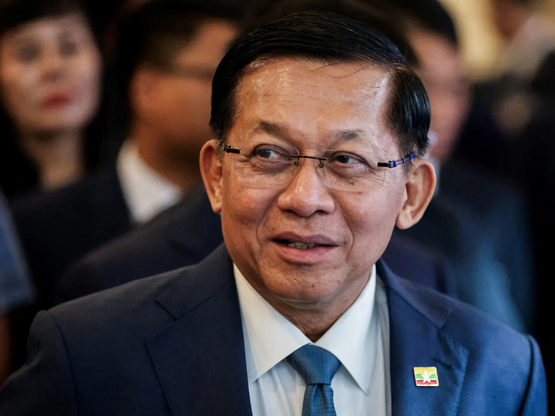 A bespectacled man in a navy suit and blue tie sits among blurred attendees.