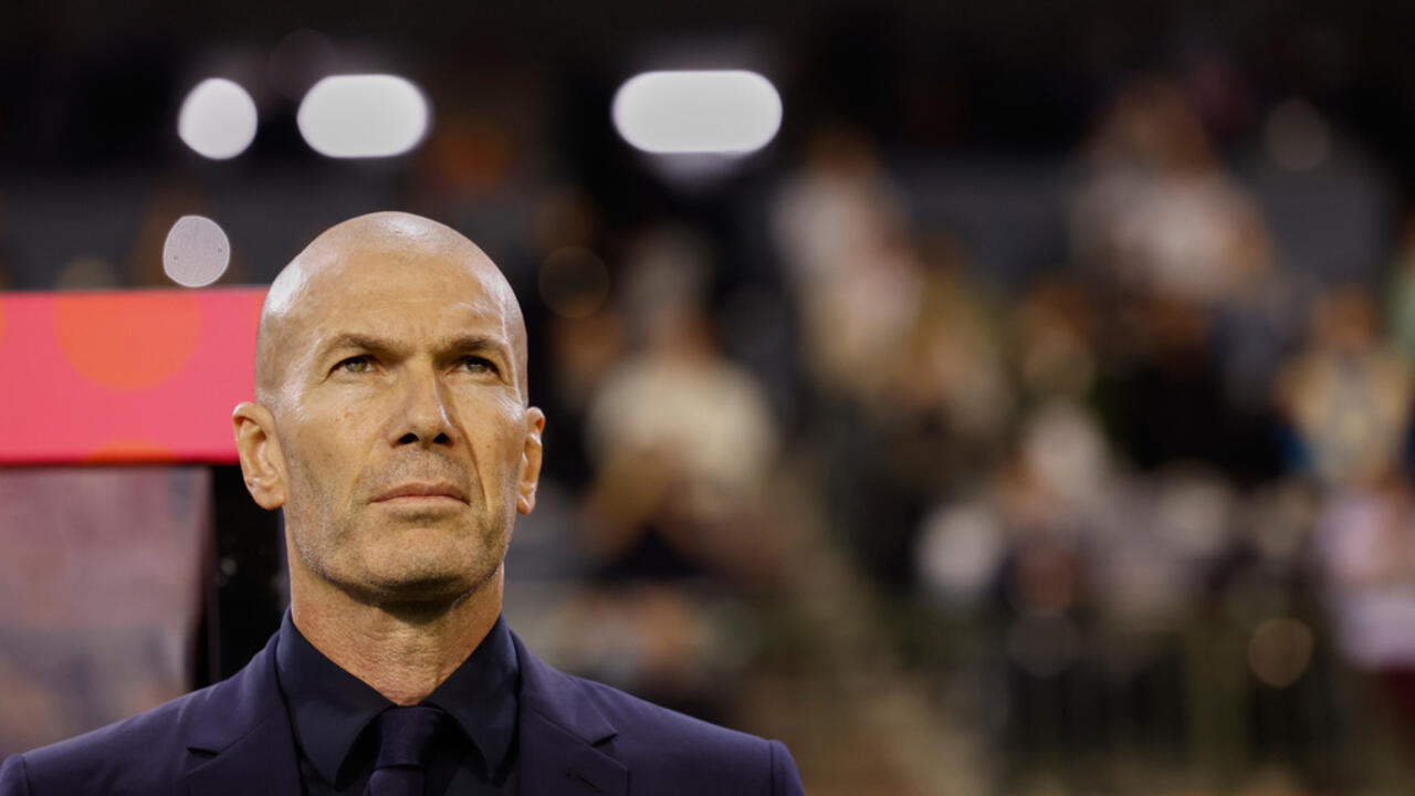 A bald man in a dark suit stands before a blurred crowd and bright stadium lights.
