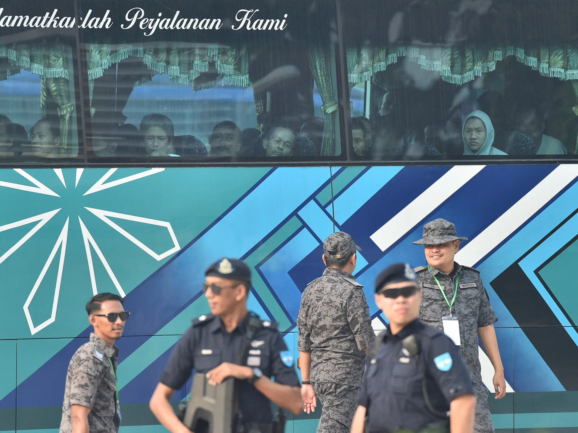 People look out from a bus as uniformed personnel stand beside its blue patterned exterior.