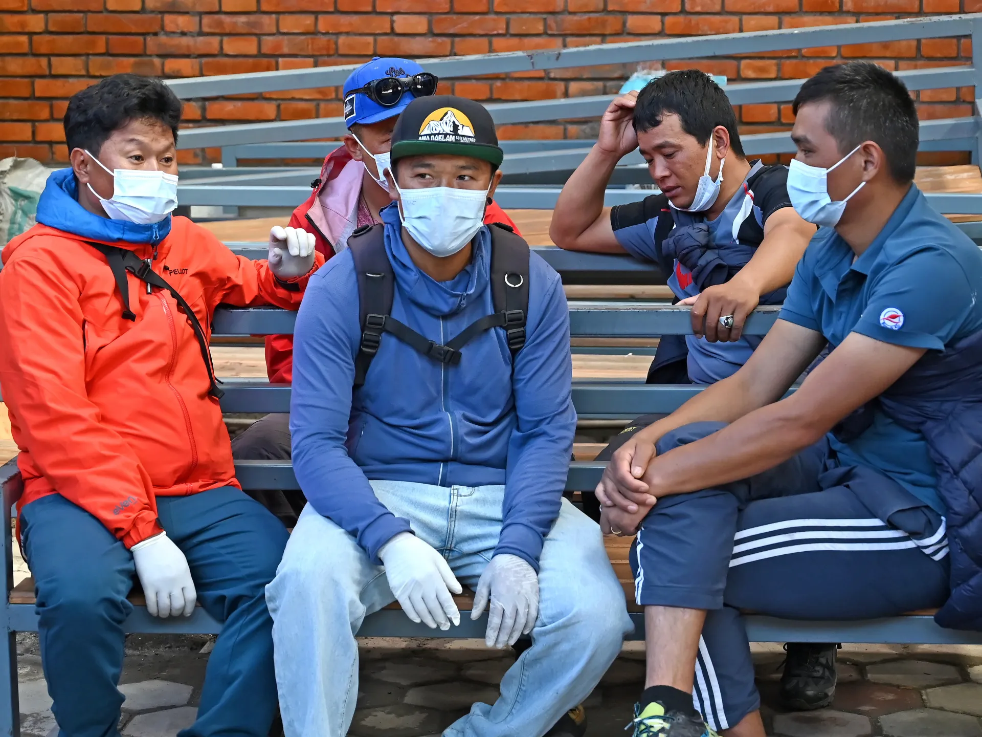 Five masked men in outdoor clothing sit together on metal bleachers, one wearing white gloves.
