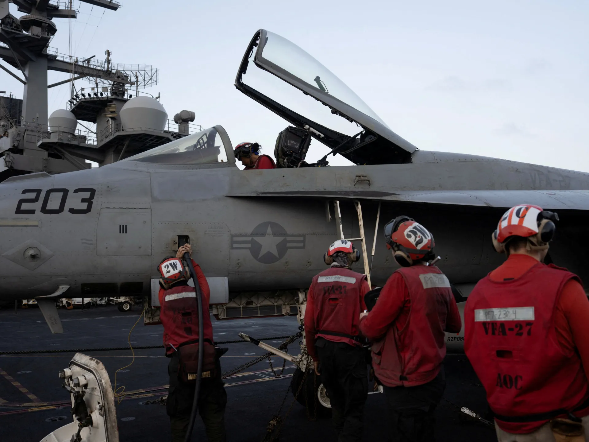 Deck crew in red vests work beside a gray fighter jet with its canopy open on an aircraft carrier.
