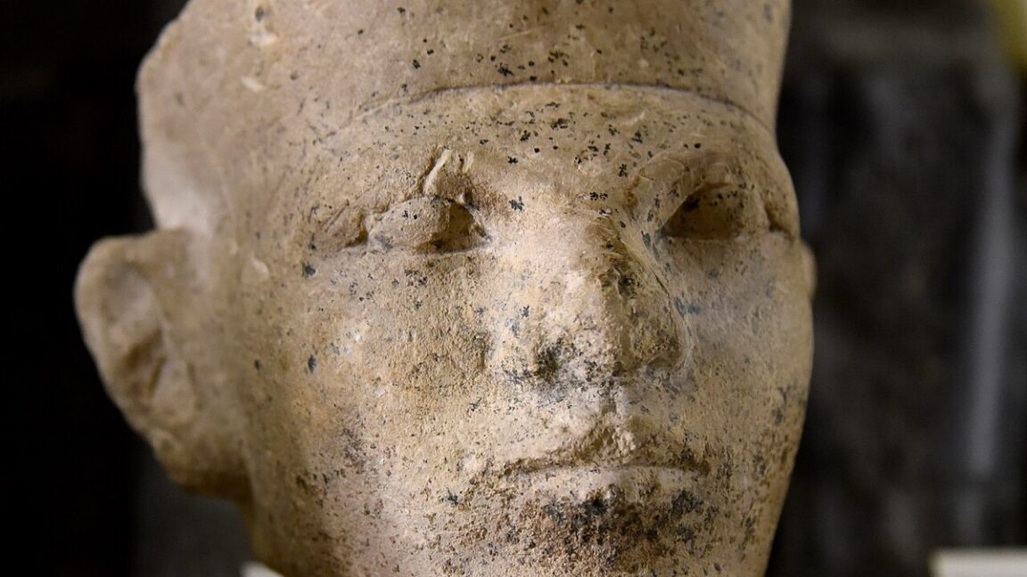 Close-up of a weathered ancient Egyptian stone sculpture's face, showing worn features and a headdress.