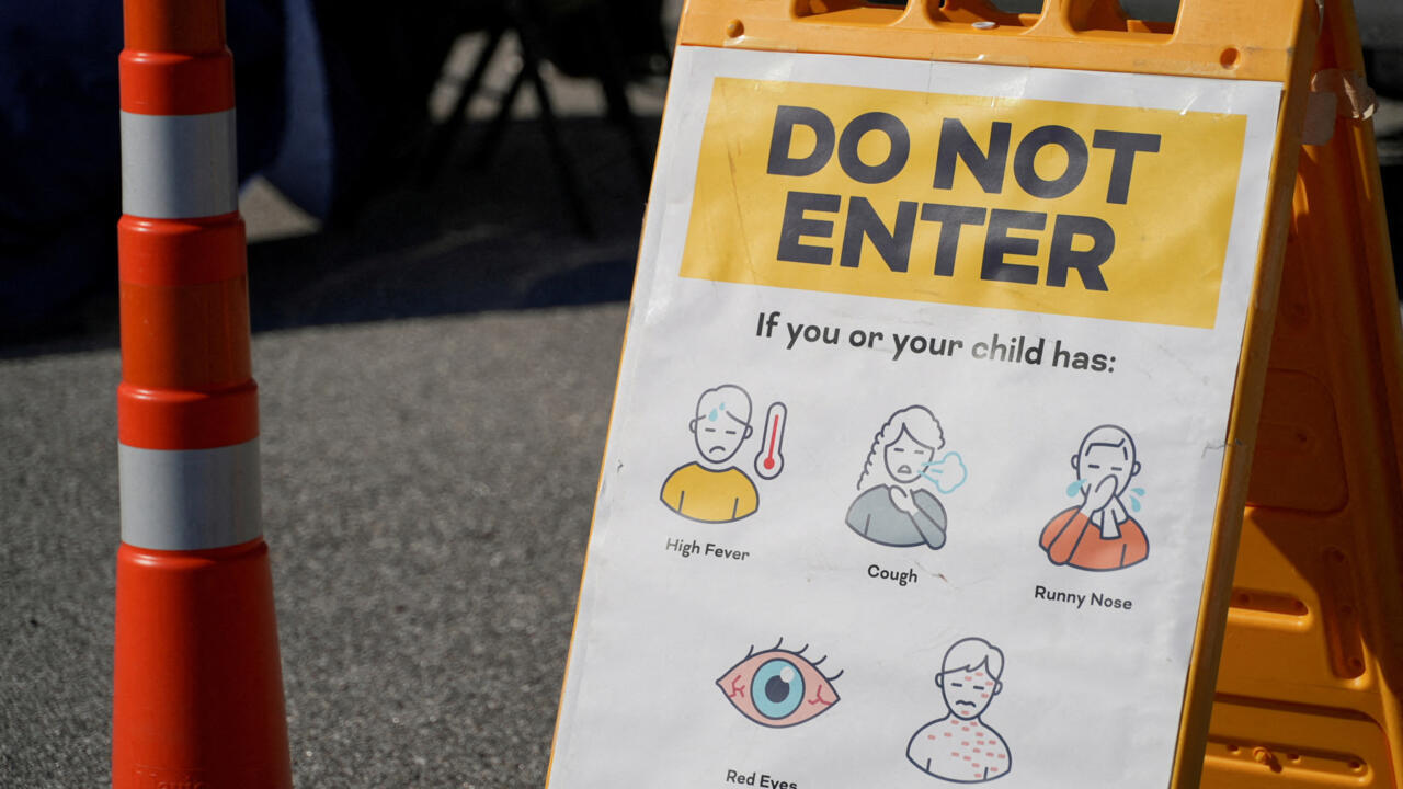 A yellow warning sign lists fever, cough, runny nose, red eyes, and rash symptoms beside a traffic cone.