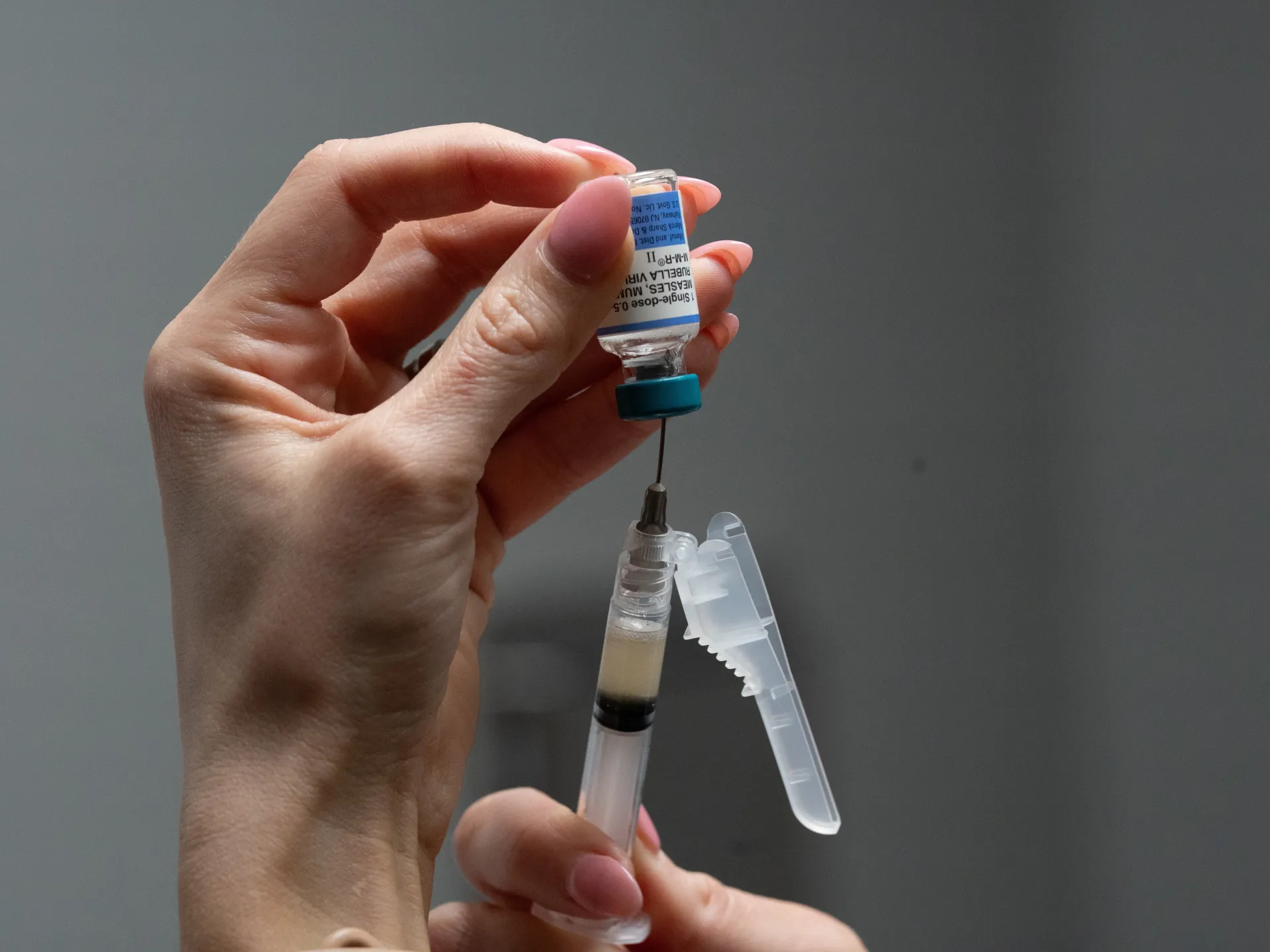 A hand holds an inverted vaccine vial above a syringe against a plain gray background.
