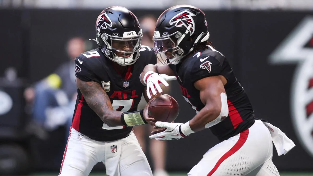 Two Atlanta Falcons players in black jerseys exchange a football during a game.