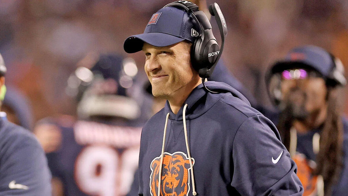 A man in a Bears hoodie, cap, and headphones stands on the sideline during a football game.
