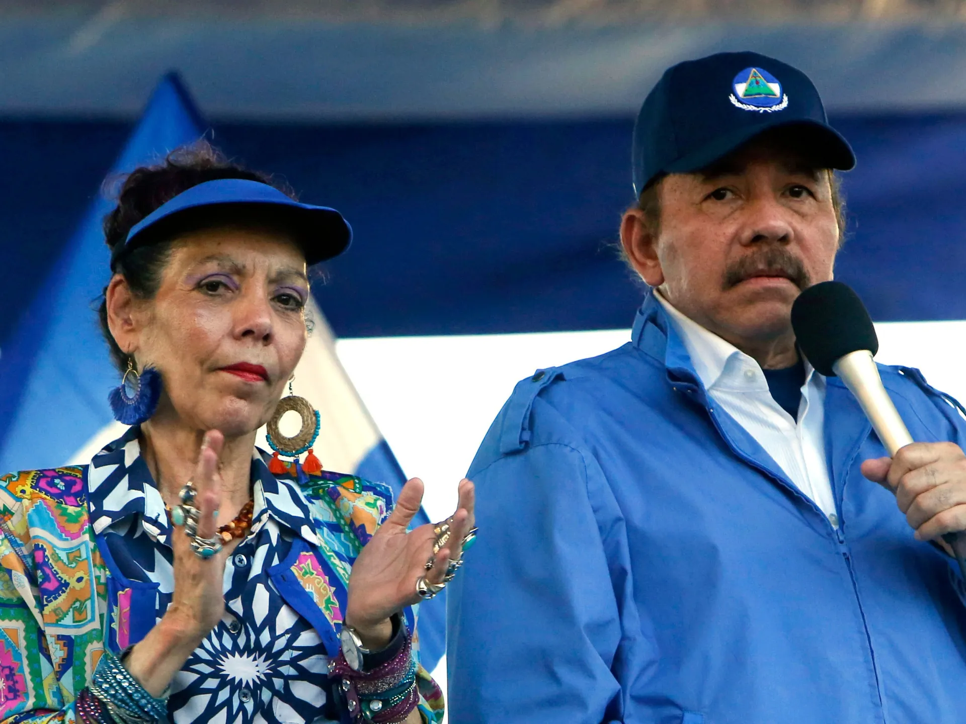 A woman in a colorful jacket claps beside a man in a blue jacket holding a microphone.