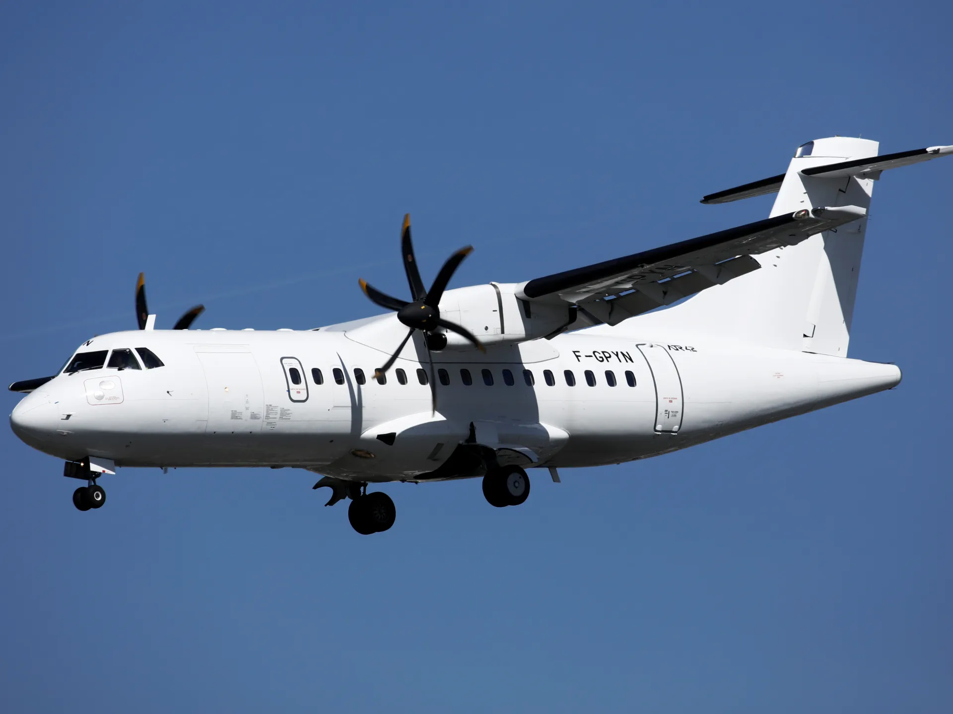 White twin turboprop passenger aircraft flies against a clear blue sky with its landing gear down.