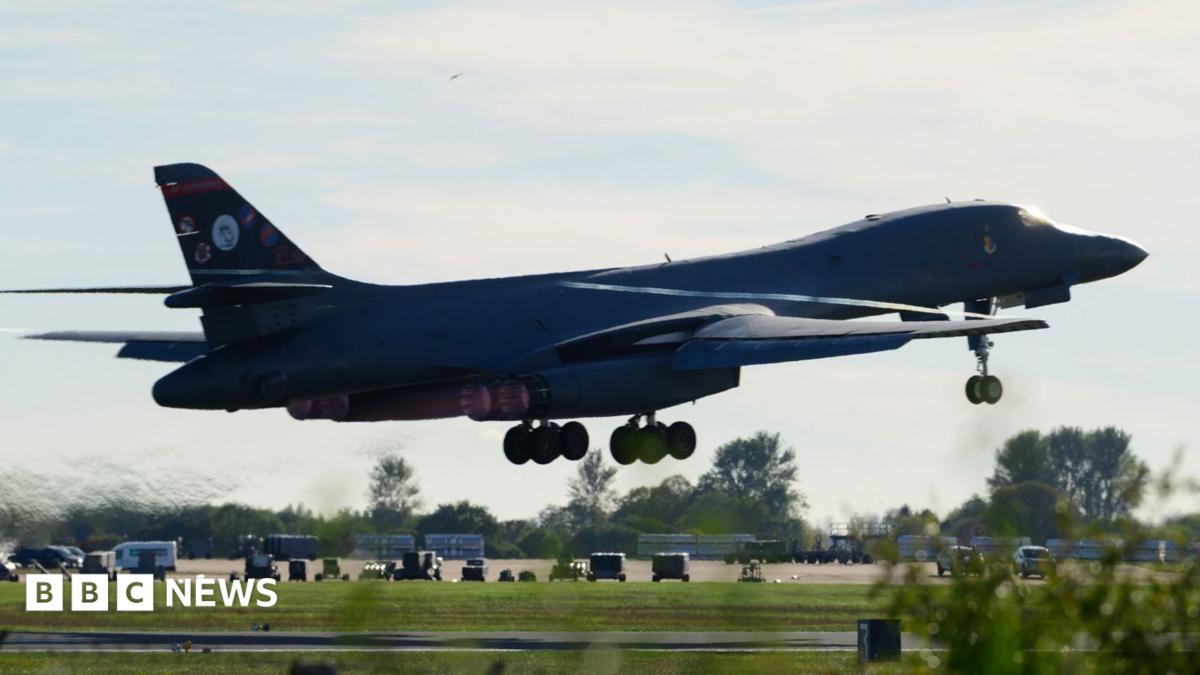 A large military bomber flies low over an airfield with its landing gear down.