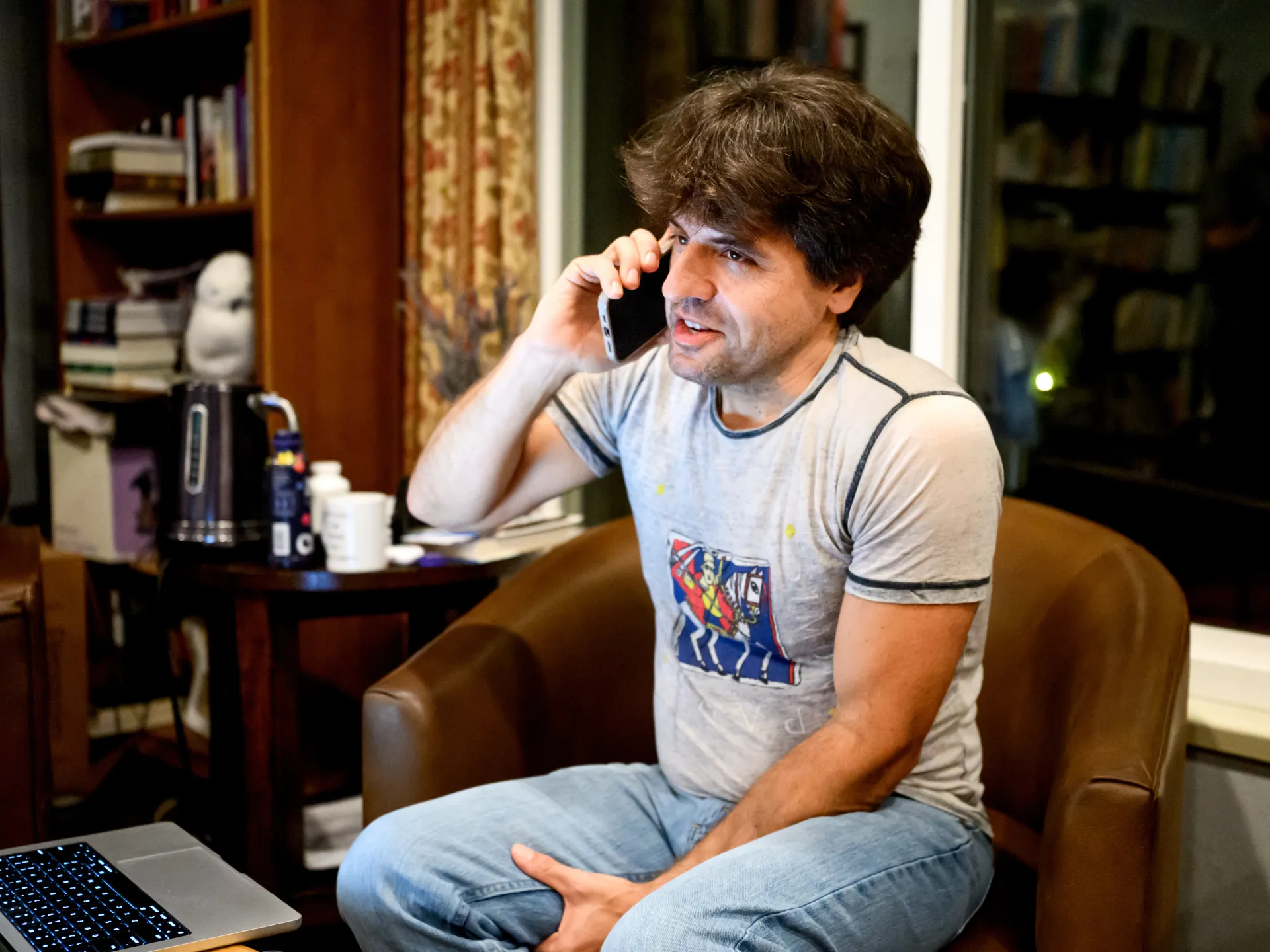 A dark-haired man sits in an armchair, holding a phone to his ear beside an open laptop.