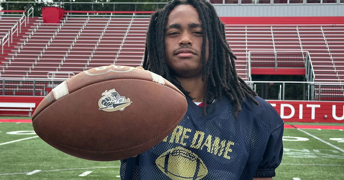 A football player holds a ball toward the camera on a field in front of bleachers.