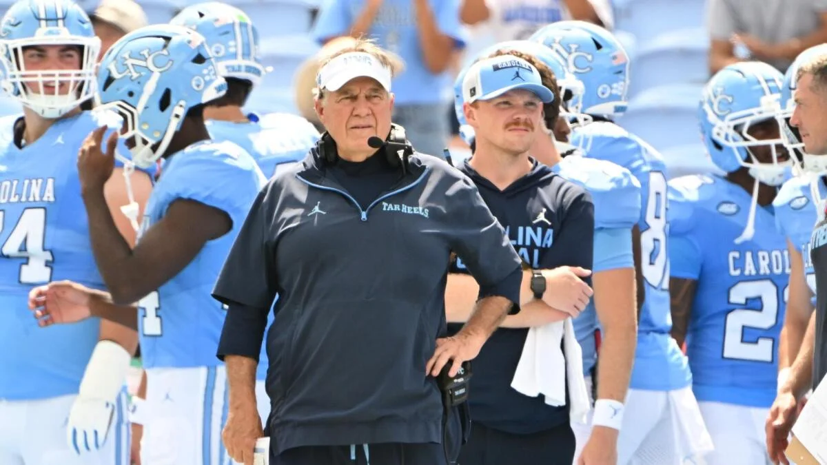 A coach stands among North Carolina Tar Heels football players in blue uniforms on the sideline.