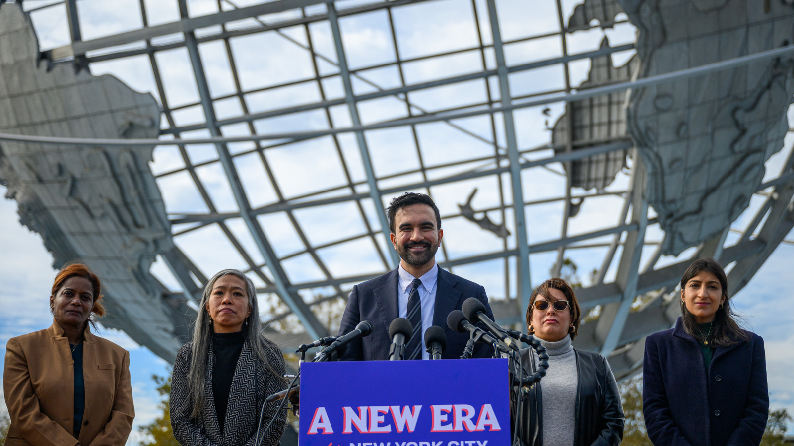 A suited man speaks at a podium surrounded by microphones and four women beneath a glass structure.