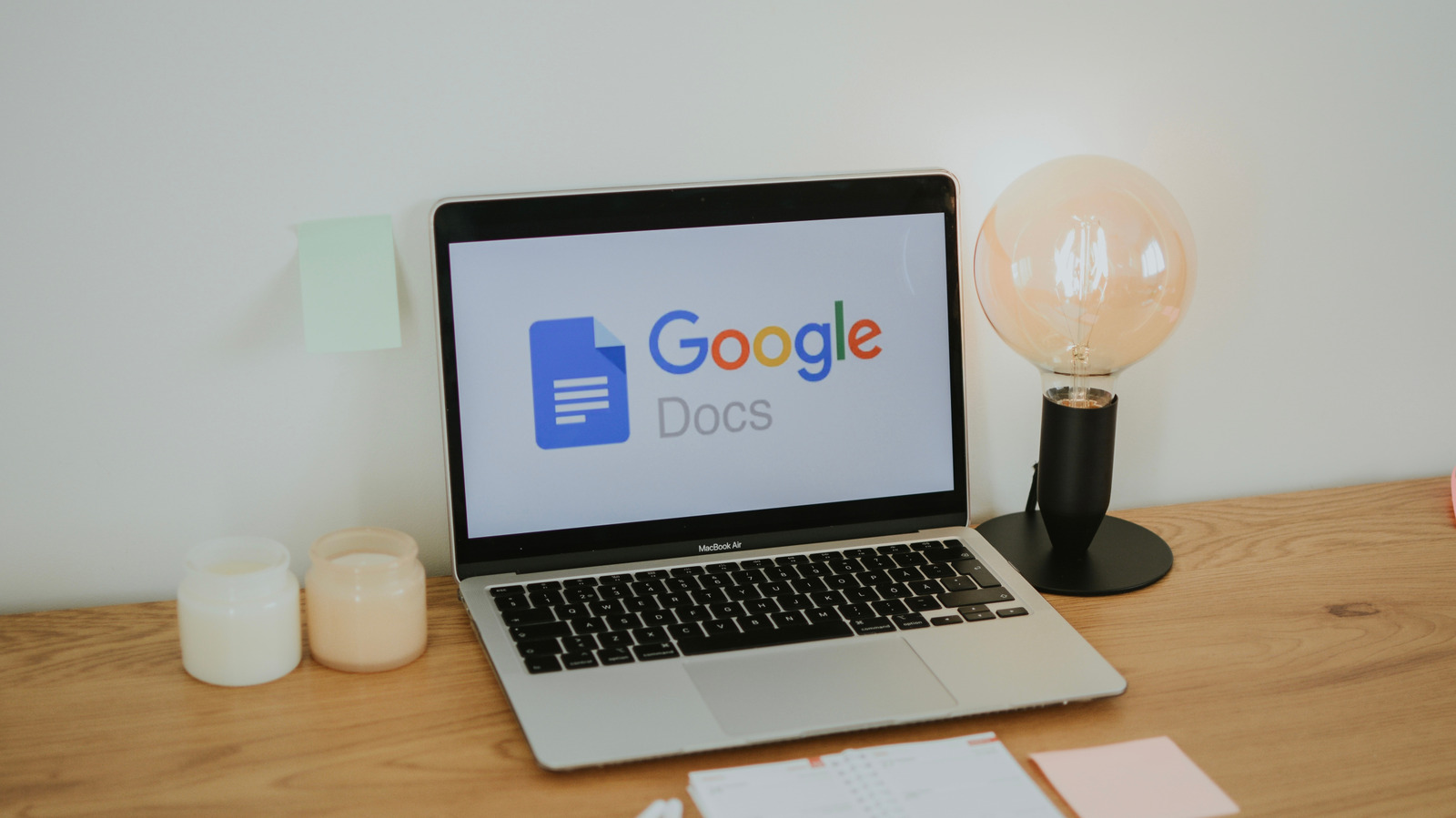 A laptop displaying the Google Docs logo sits on a wooden desk beside a lit bulb.