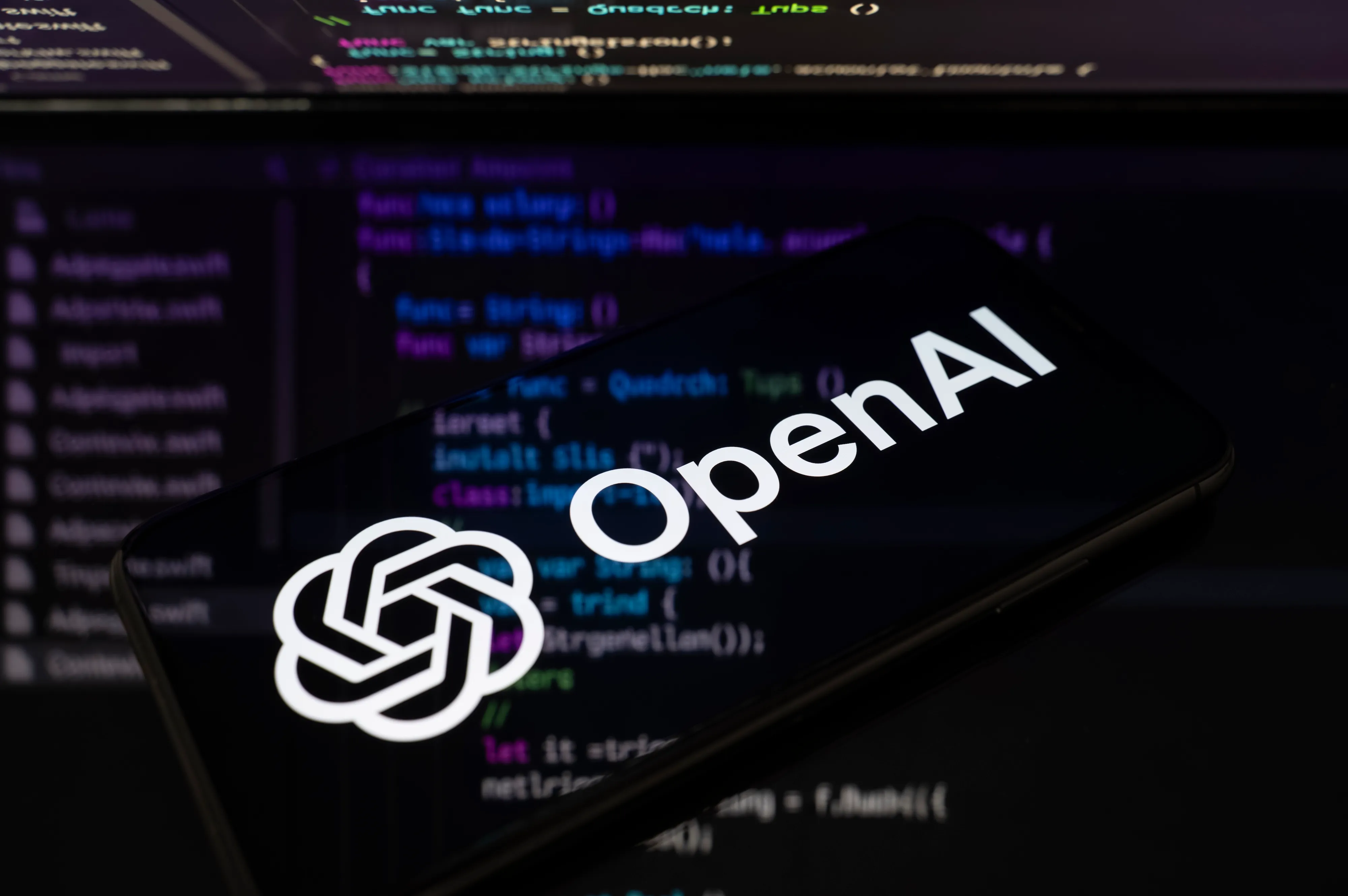 A smartphone displaying the OpenAI logo rests in front of a blurred computer screen showing code.