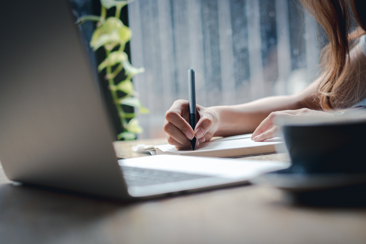 A person writes in a notebook beside an open laptop, with a leafy plant nearby.