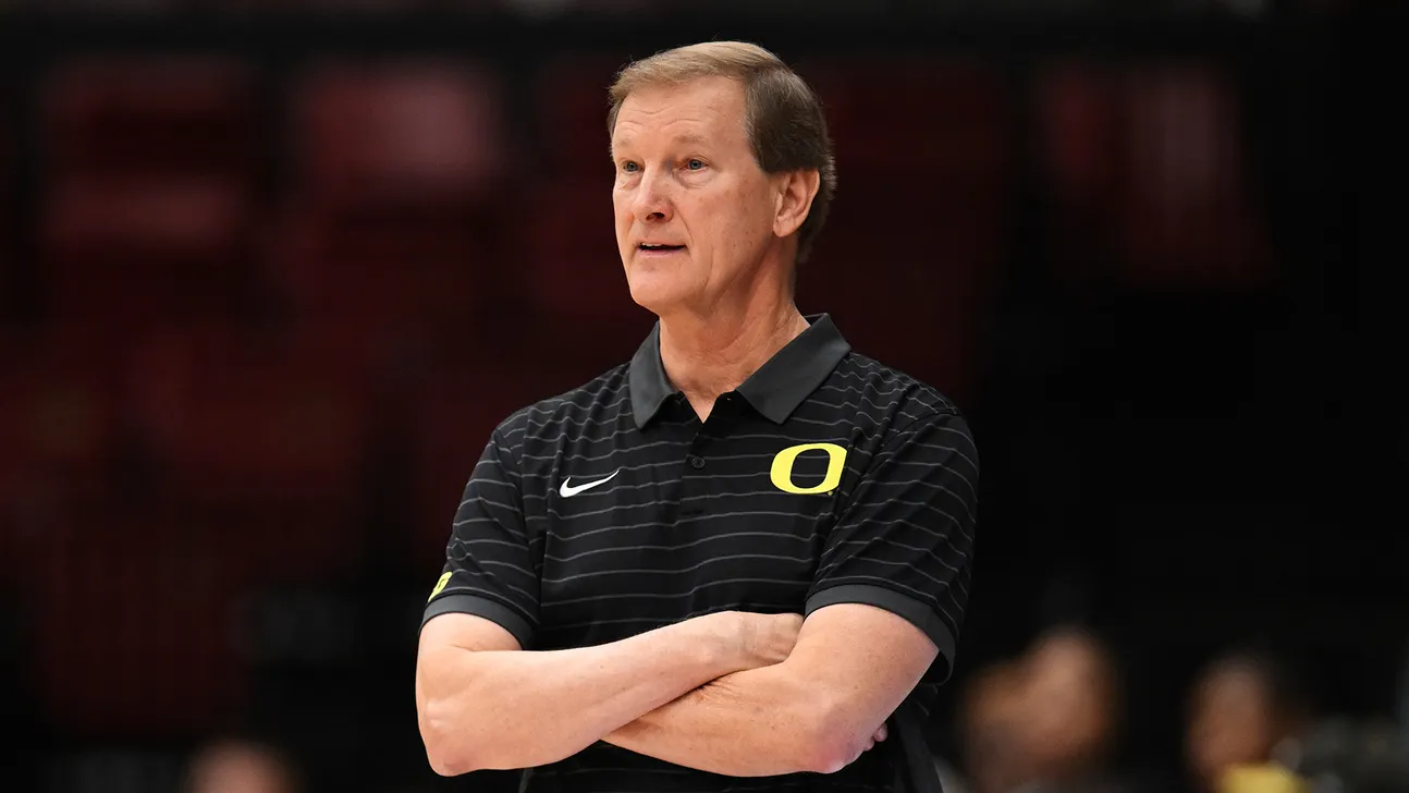 A man in a black Oregon polo stands with arms crossed in a basketball arena.