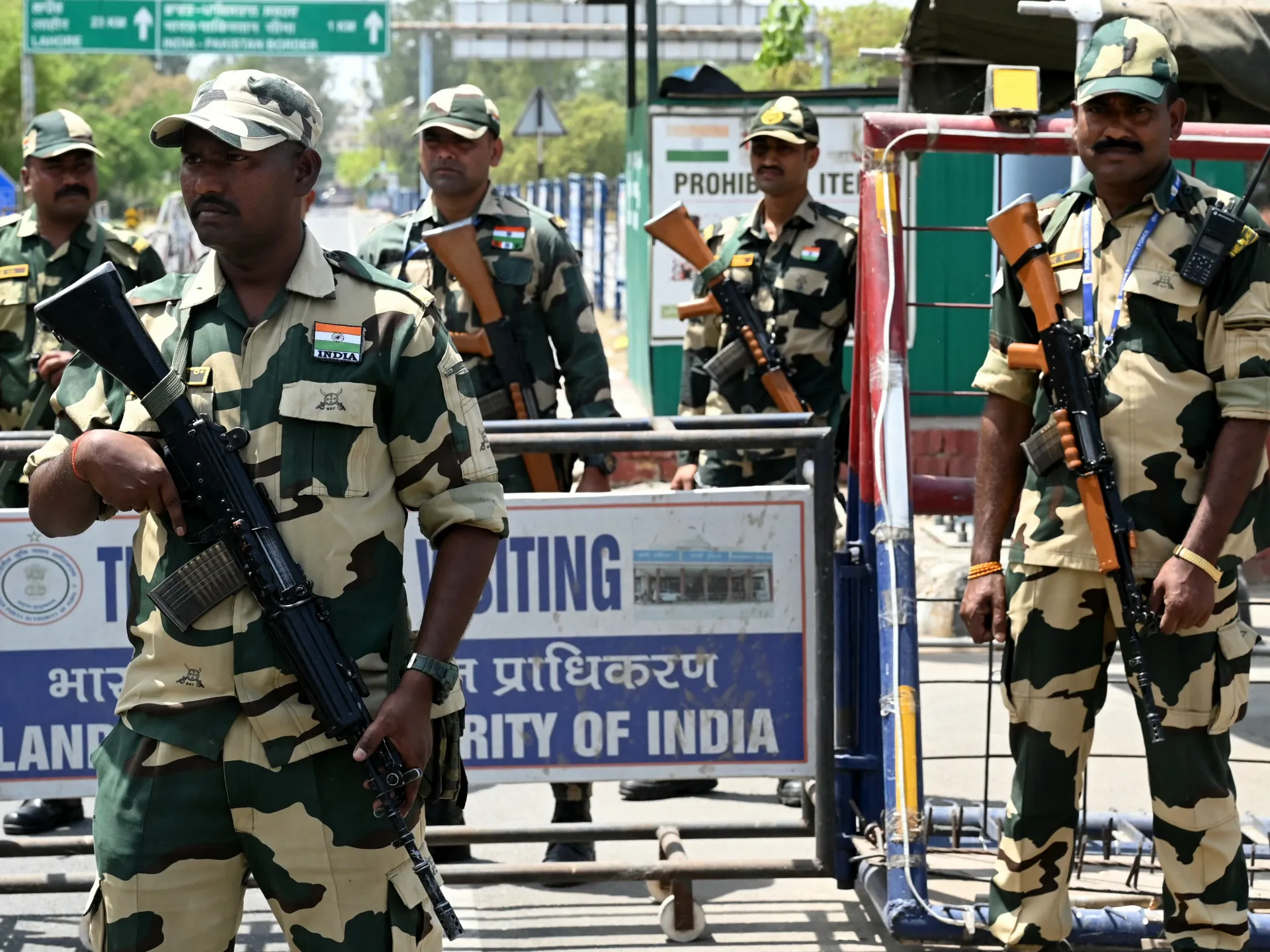 Armed personnel in camouflage uniforms stand at a security checkpoint beside barricades and an authority sign.