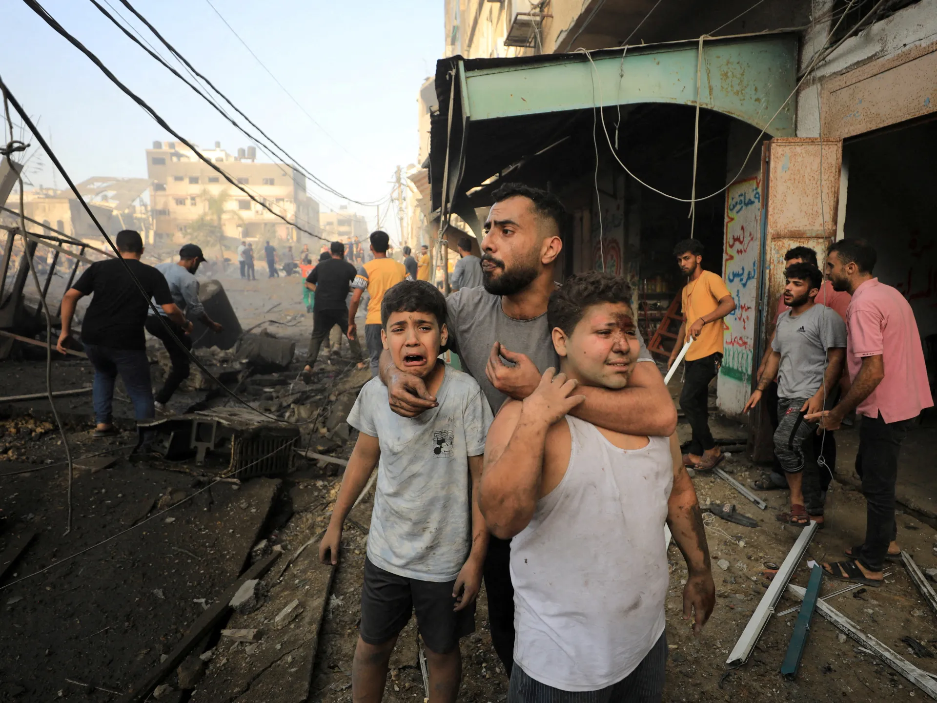 A man comforts two distressed boys amid rubble and damaged buildings as people gather in a debris-strewn street.