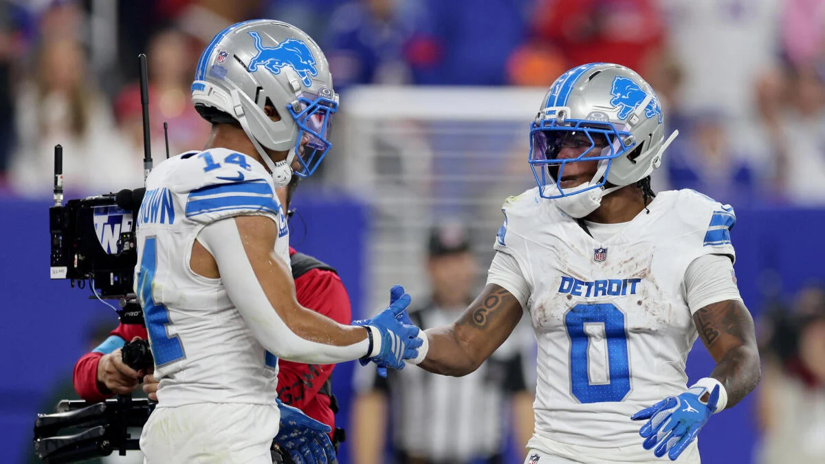 Two Detroit Lions players in white uniforms and silver helmets clasp hands on the field.