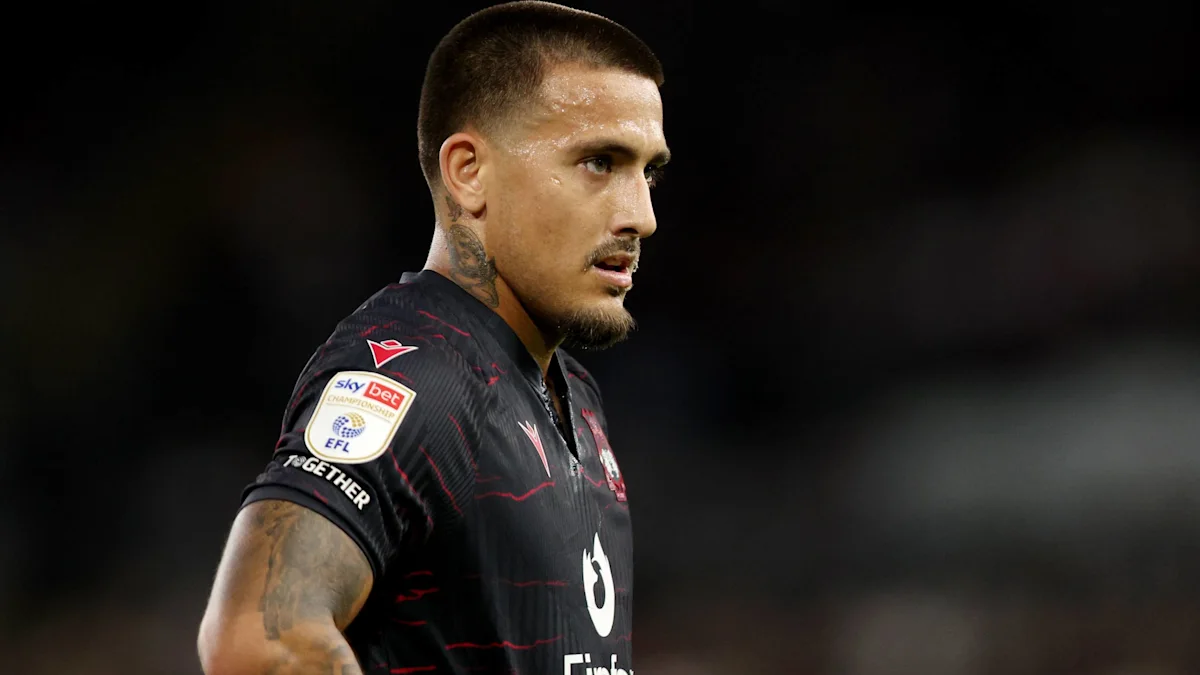 Footballer in a black jersey with red detailing looks to the side under stadium lights.