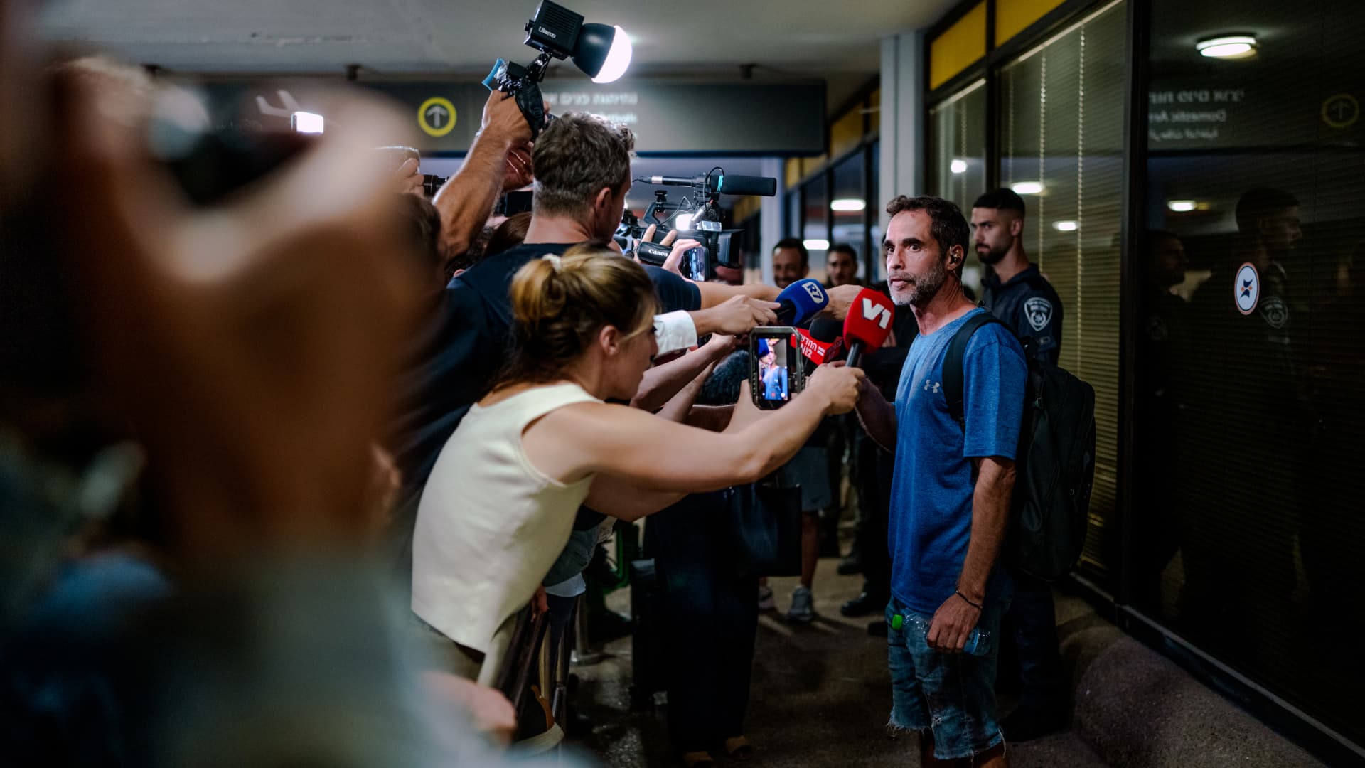 A man in a blue shirt speaks with reporters and cameras in a crowded indoor corridor.