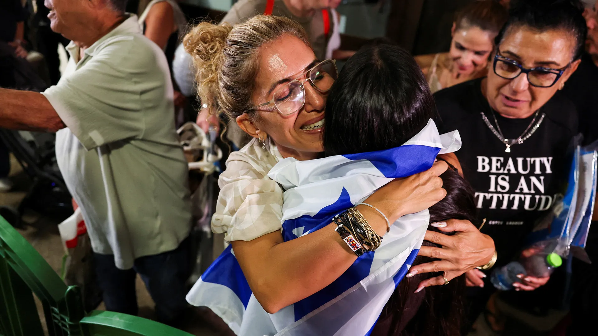 Two women embrace, one draped in an Israeli flag, as another woman watches nearby.