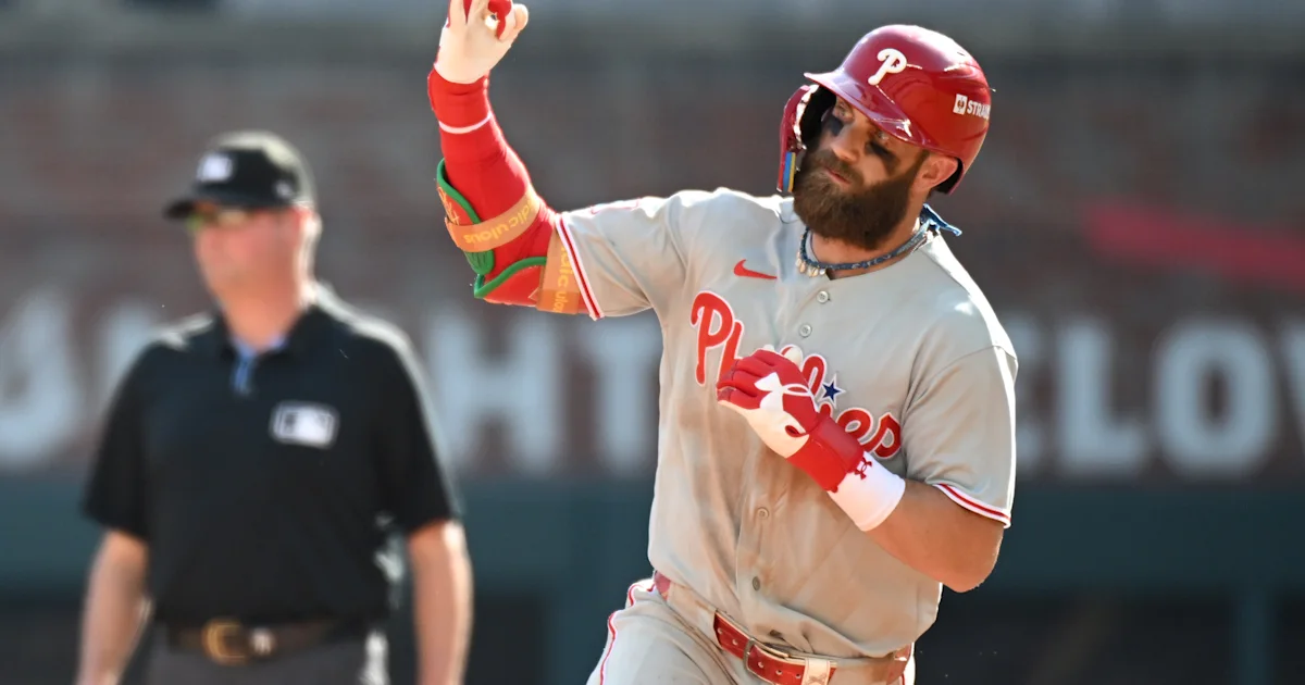 Bearded Phillies player in gray uniform raises one hand while standing near a blurred umpire.