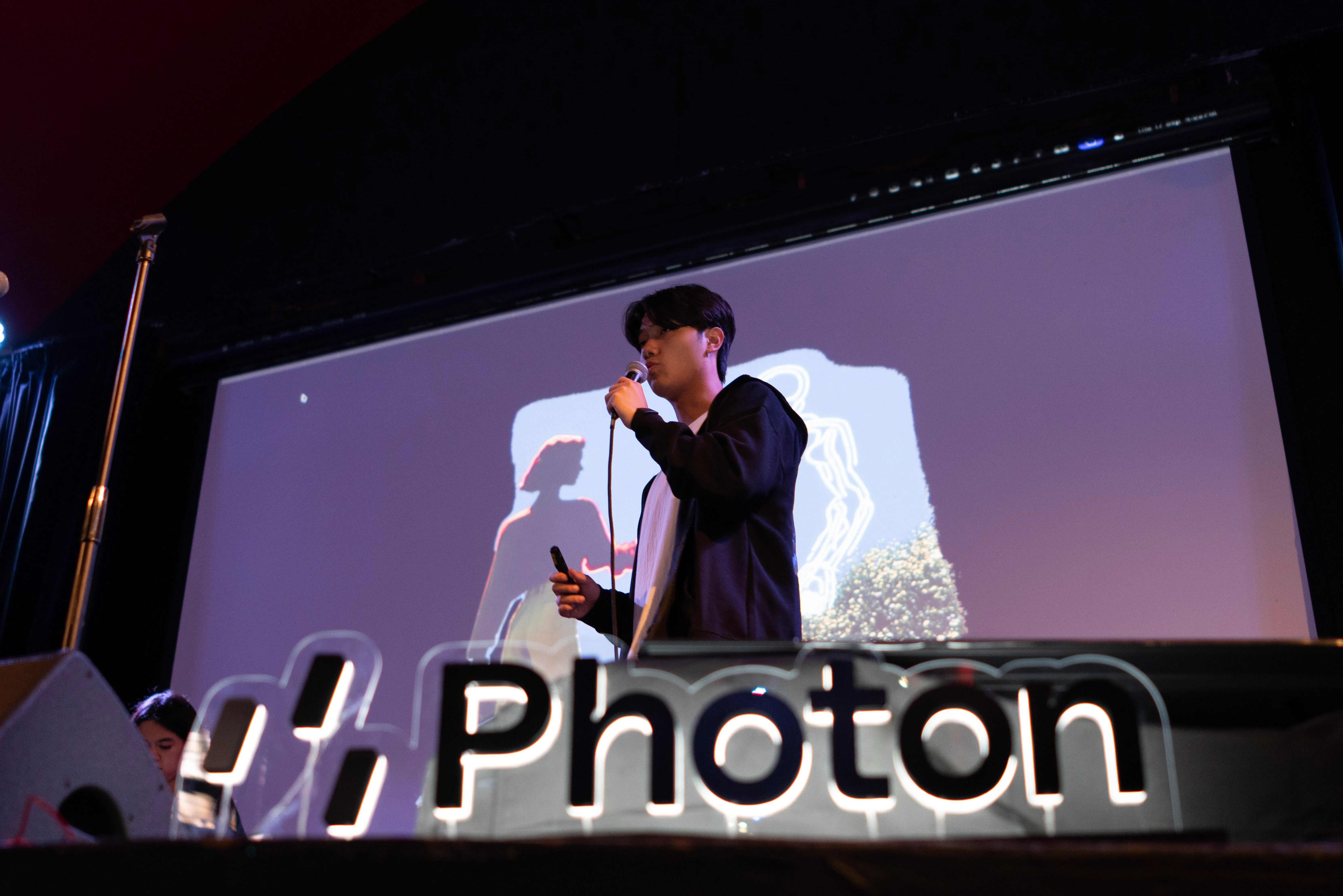 A man speaks into a microphone onstage before a projected image and large Photon lettering.