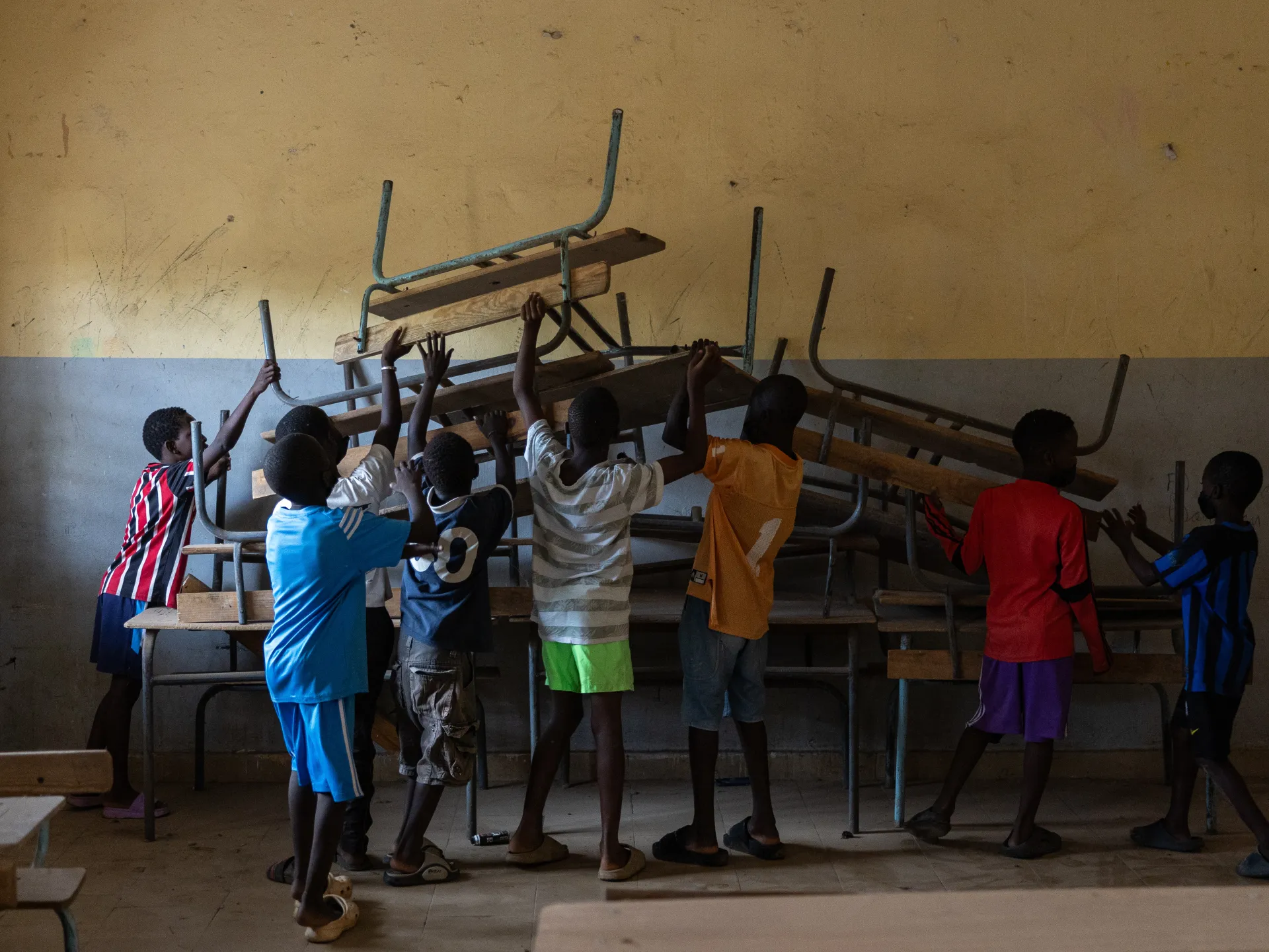 Children lift and move stacked wooden desks and metal frames inside a worn classroom.