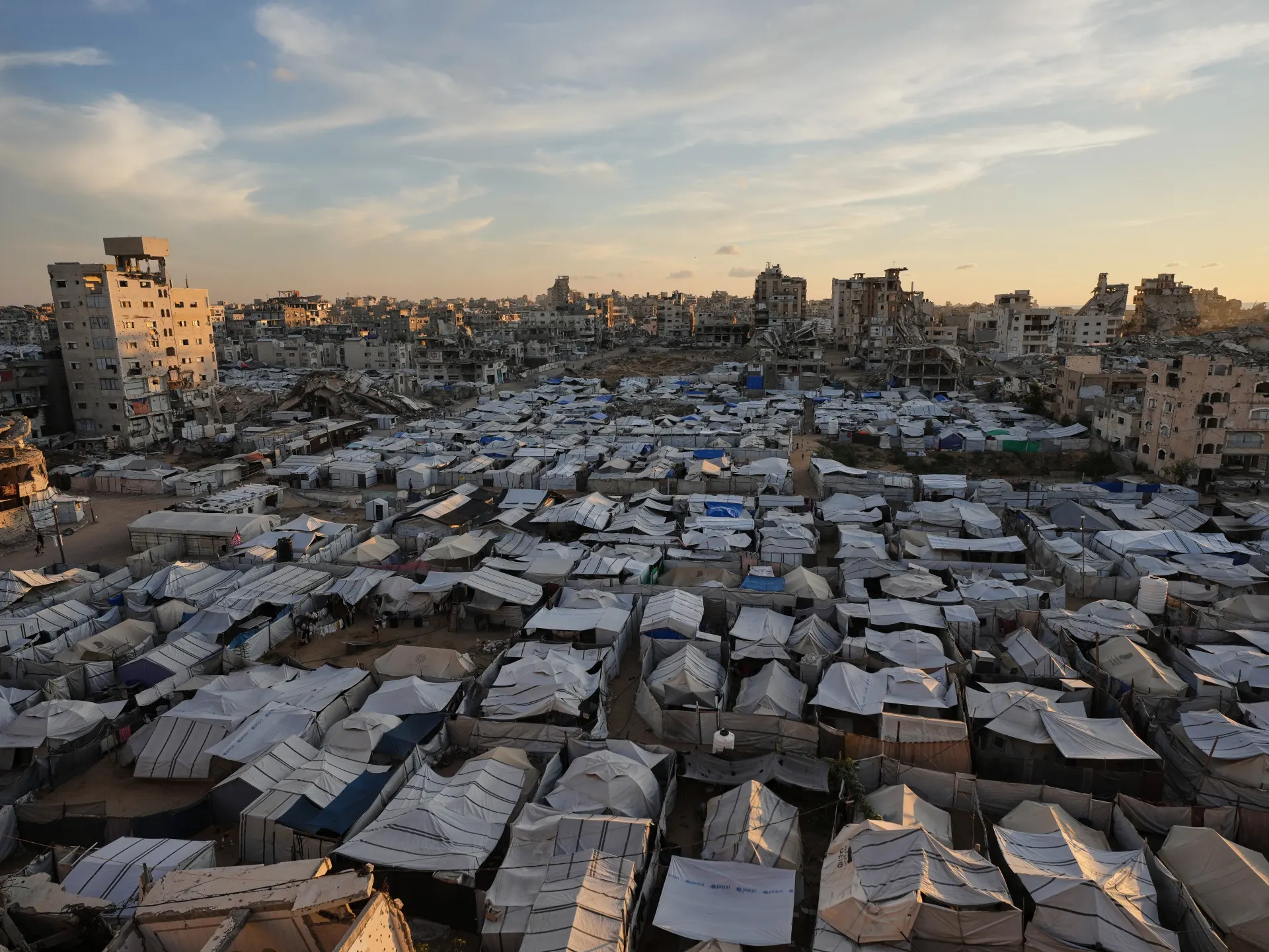 Dense tent camp fills the foreground beneath a skyline of damaged buildings at sunset.