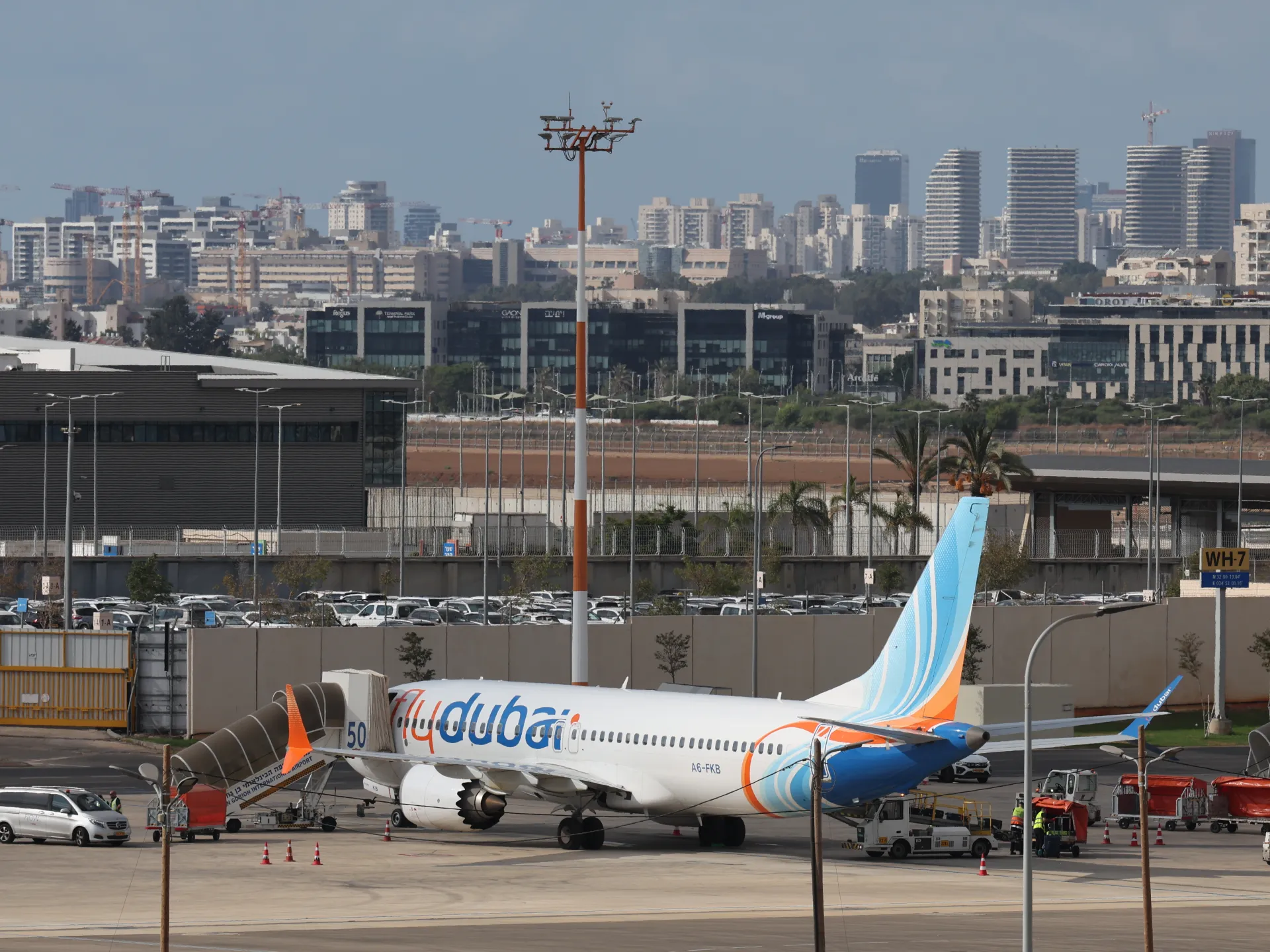 A Flydubai passenger jet sits on an airport apron, with service vehicles and city buildings behind it.