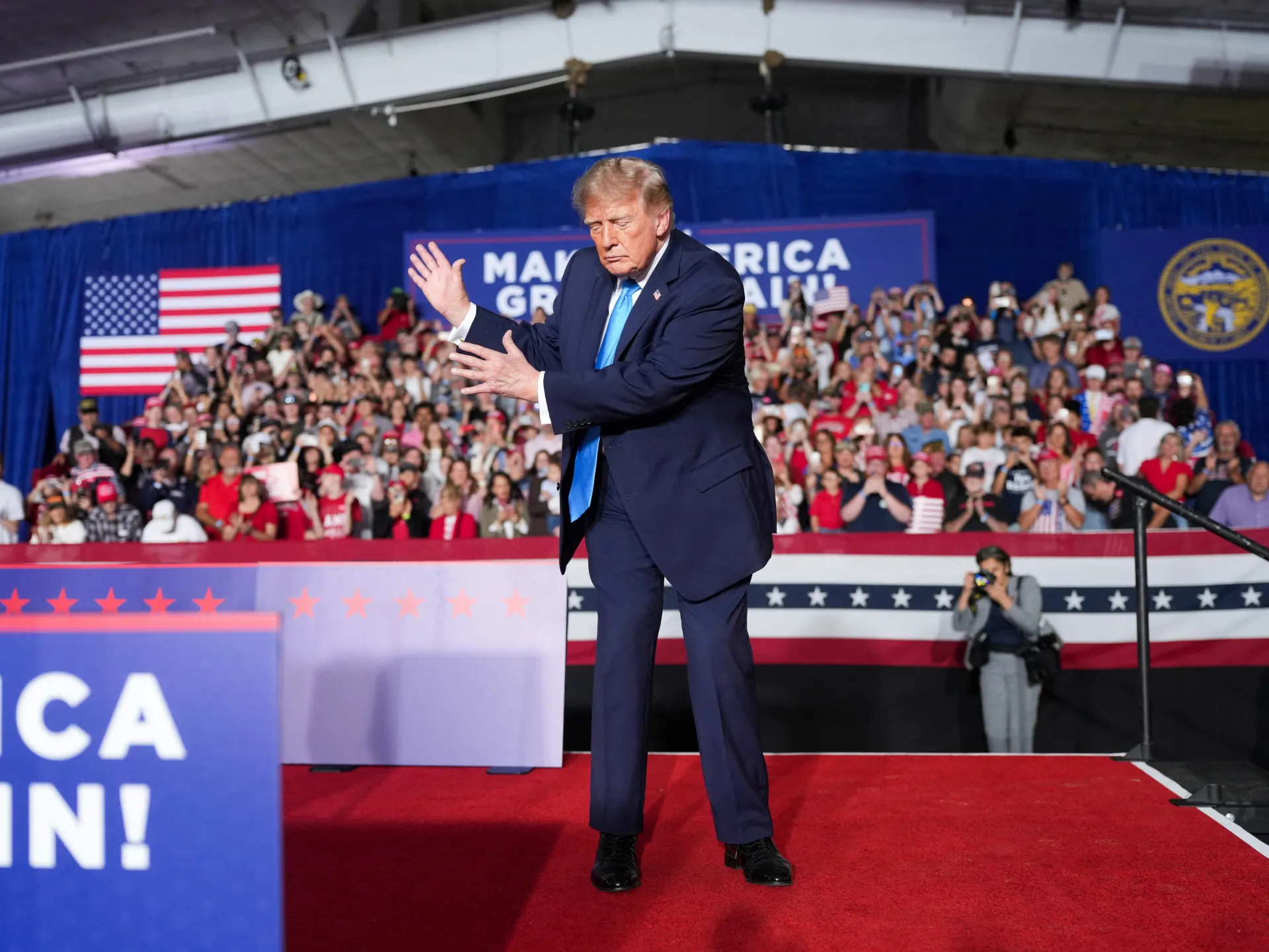 A suited man gestures on a red-carpeted stage before a crowd at a political rally.