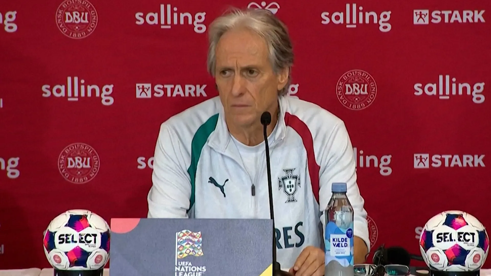 An older man in a white Portugal team jacket sits at a press table with a microphone, footballs and water bottle.