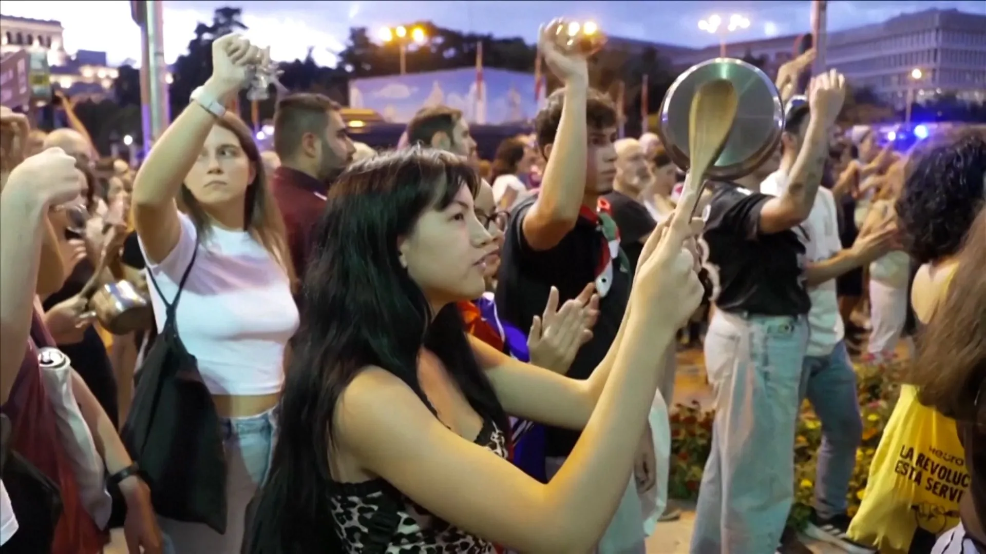 People gather outdoors at dusk, raising pots and pans and clapping among a crowd.