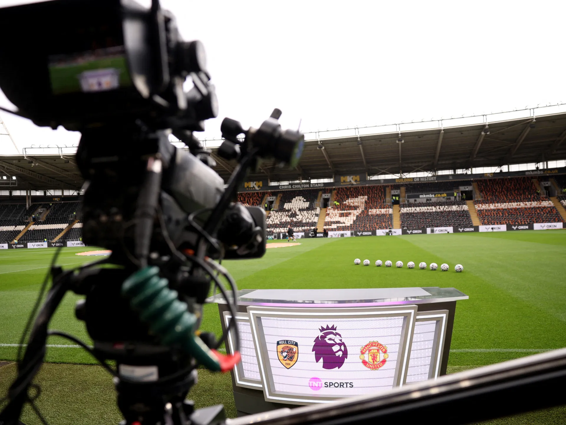A broadcast camera overlooks a football pitch, lined balls, and a studio desk with team and league logos.