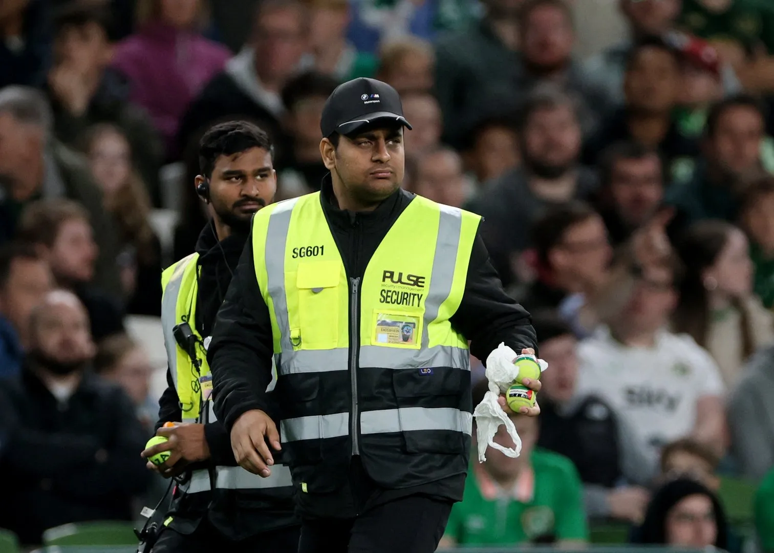 A stadium security guard in a fluorescent vest holds tennis balls and a white cloth as spectators watch.