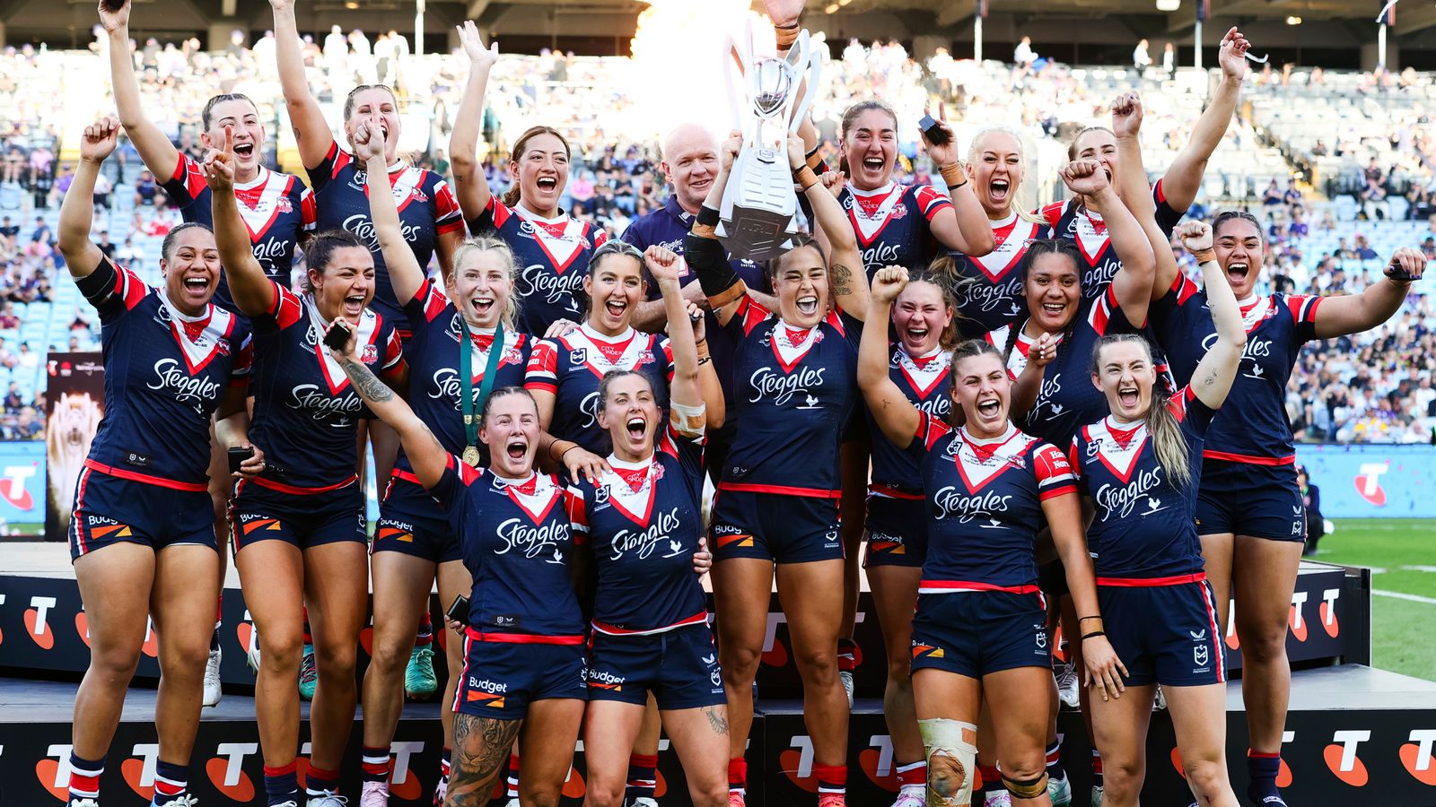 Roosters women’s rugby league players celebrate together, raising their arms around a trophy on the field.