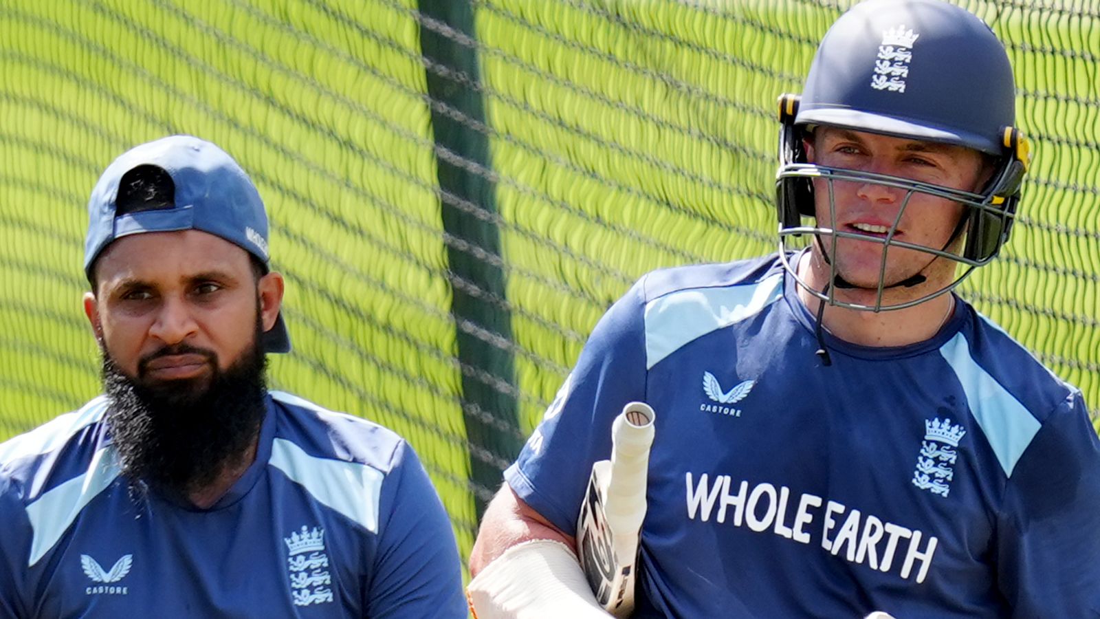 Two England cricketers in blue training kits stand beside a practice net, one wearing a helmet and holding a bat.