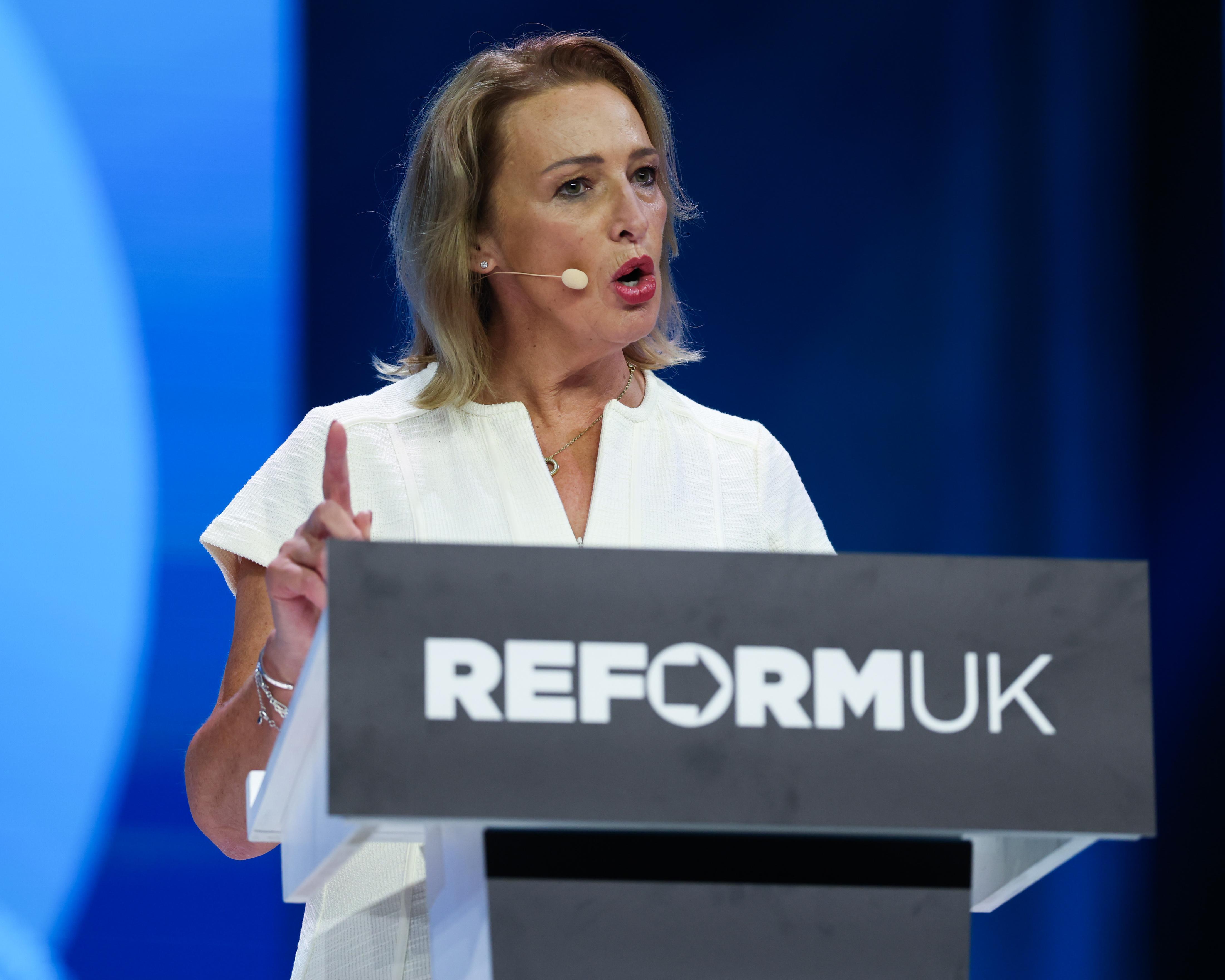 A woman speaks at a Reform UK lectern, raising one finger against a blue stage backdrop.
